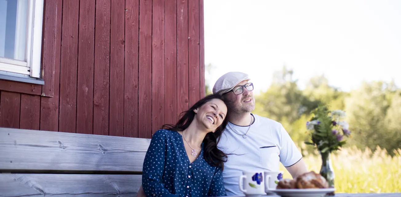 Par som sitter och fikar framför röd stuga.